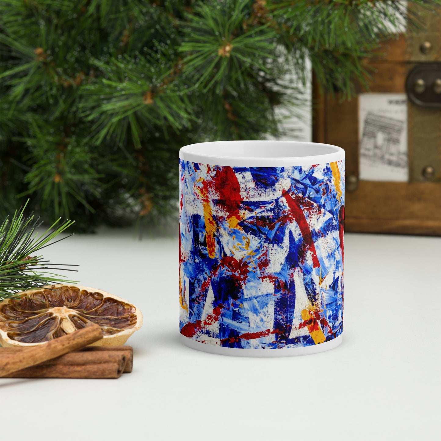 Ceramic mug featuring the abstract artwork "Rebel Parade" with bold red, blue, and white brushstrokes wrapping around the surface, shown with branches, foliage and decor